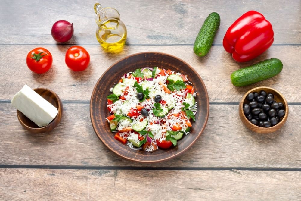 Salata bulgareasca in bol din ceramica alaturi de legume si branza pe masa rustica